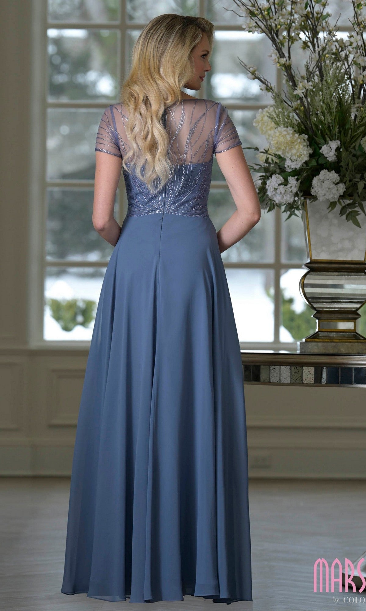 A woman with long blond hair wears the Marsoni M351 long blue chiffon A-line formal dress, floor-length and short-sleeved, standing indoors by a window and a vase of white flowers.